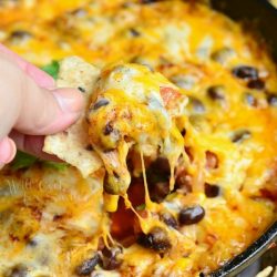 a black skillet with hot 7 layer dip rests on a table surrounded by chips and a hand dipping one chip into the dip