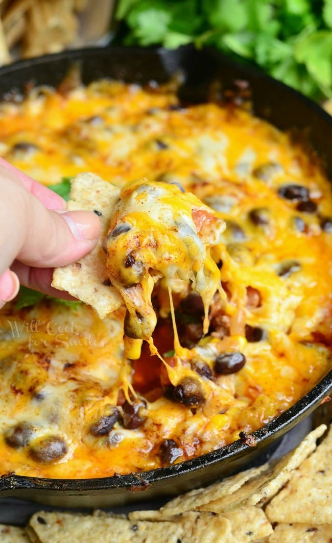 dipping a chip into the Hot 7 Layer Dip with cheese and black beans in a cast iron pan 