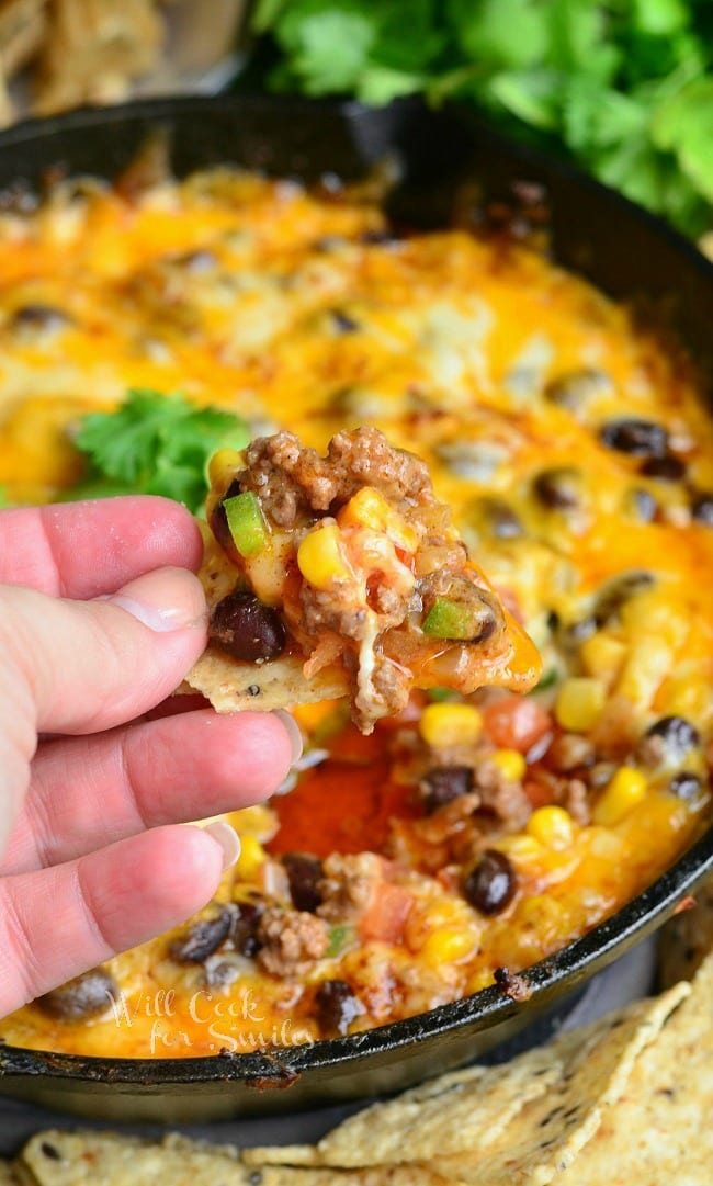 a black skillet with hot 7 layer dip rests on a table surrounded by chips and a hand dipping one chip into the dip