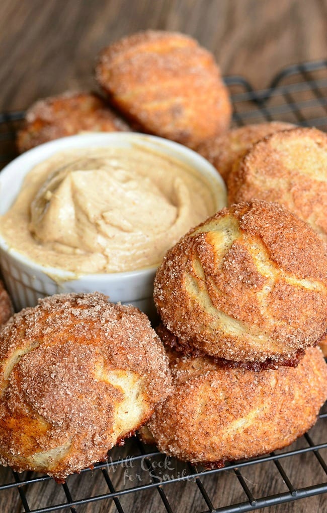 Snickerdoodle Pretzel Puffs served on a wire rack around a small, white dish containing a creamy cinnamon dip. These soft, fluffy little pretzel buns are made and coated with cinnamon and sugar.