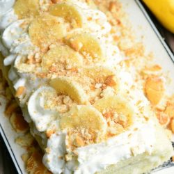 view from above of banana pudding cake roll on a decorative white rectangular plate on a wooden table with 2 bananas in the background