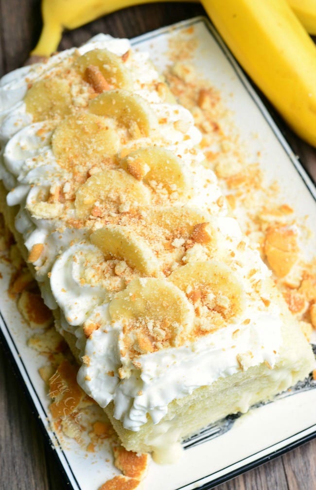 view from above of banana pudding cake roll on a decorative white rectangular plate on a wooden table with 2 bananas in the background