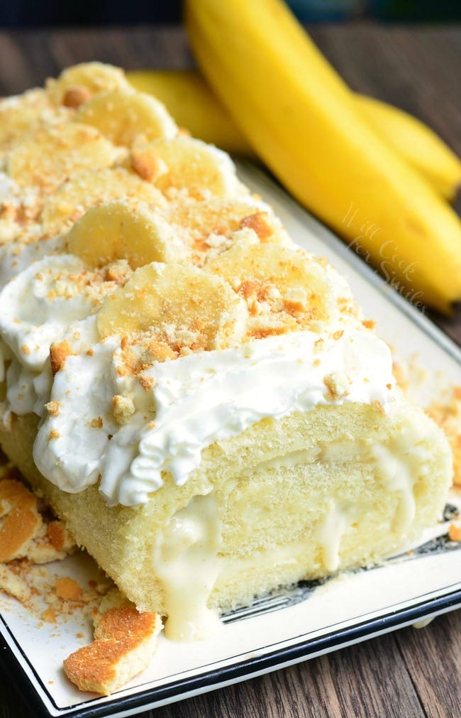 Banana Pudding Cake Roll on a plate with whipped cream, crushed vanilla waffers and banana on top with a unpeeled banana to the right  