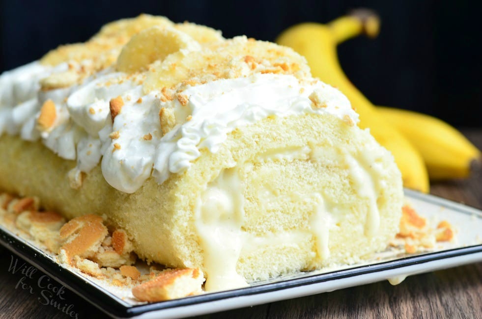 horizonal photo of Banana Pudding Cake Roll on a plate with whipped cream, crushed vanilla waffers and banana on top with a unpeeled banana to the right  