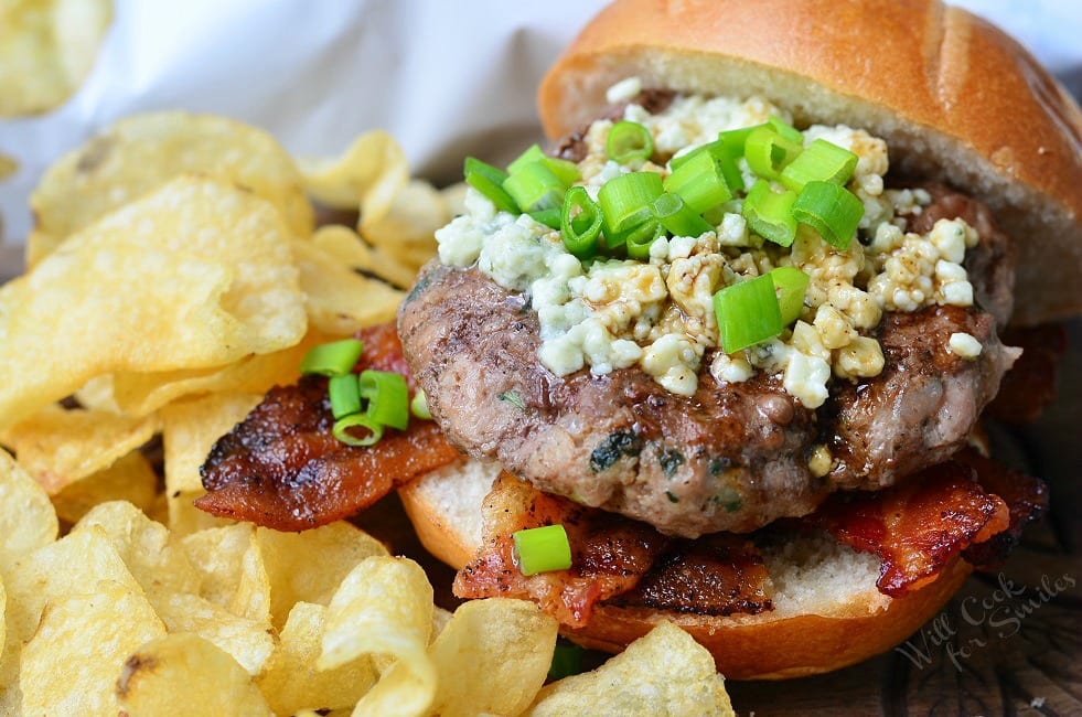 Bacon Burger on bun with blue cheese on top with green onions on top in a bun with regular chips around it 