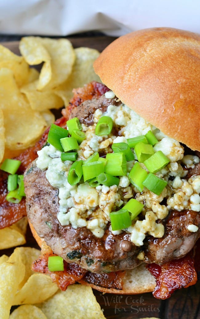 Bacon Burger on bun with blue cheese on top with green onions on top in a bun with regular chips around it 