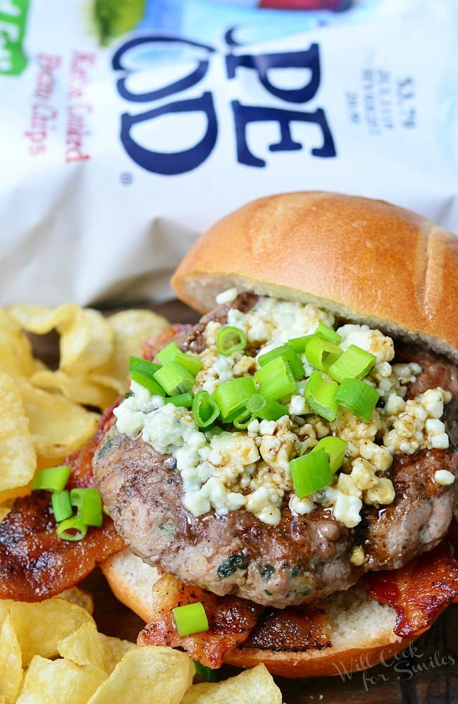 Bacon Burger on bun with blue cheese on top with green onions on top in a bun with regular chips around it 