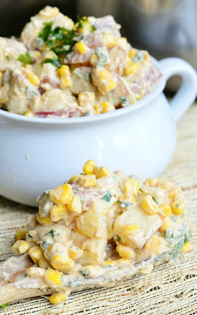 Chipotle Ranch Potato Salad with potatoes, corn, and parsley on top  in a white bowl and a wooden spoon in front of the bowl with the potato salad 