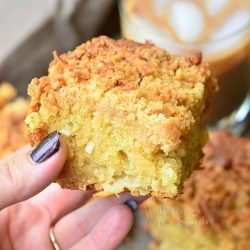 2 portions of coconut coffee cake on a stone table in front of a glass of iced coffee and a hand holding 1 portion above