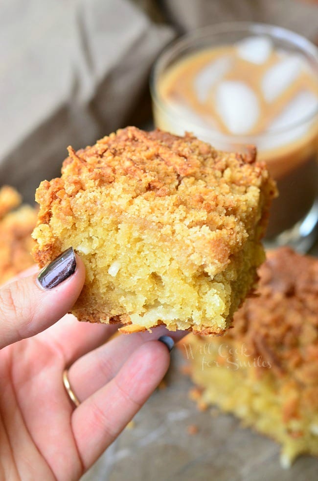A piece of Coconut Coffee Cake is held in the left hand for a close up. The rest of the cake and an iced coffee are in the background.