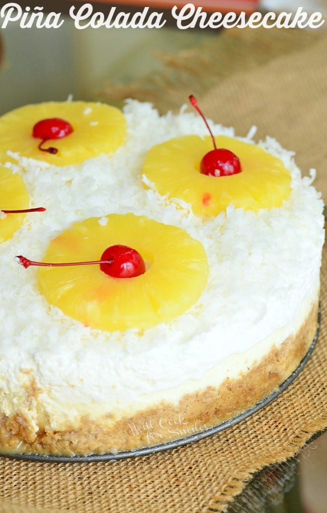 Pina Colada Cheesecake sits on the bottom pan. The top of the cheesecake is covered in white coconut flakes. Several yellow pineapple rings are placed on top with a red cherry in the middle of each hole.