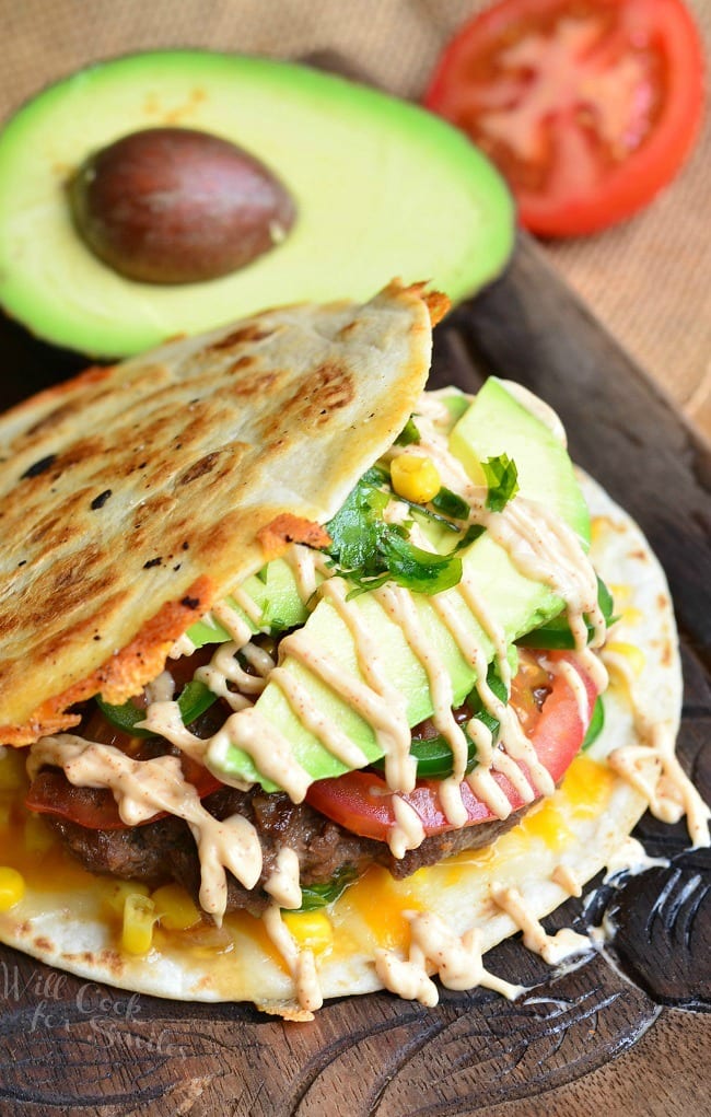 Quesadilla Burger with avocado tomato and fried tortilla as the bun with half of a avocado and a half of a tomato in the background 