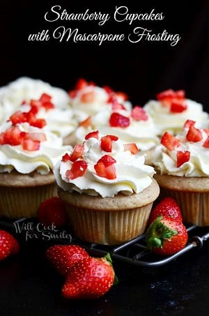 Strawberry cupcakes with white frosting and cut up strawberries on top on a cooling rack with strawberries around it 