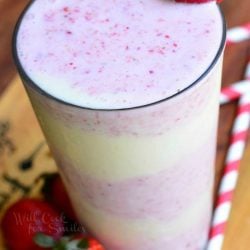 view from above of pint glass filled with strawberry shortcake milkshake on a wooden table with strawberries and red and white striped straws scattered at bottom of glass