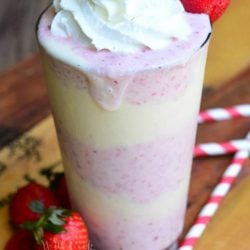 pint glass filled with strawberry shortcake milkshake on a wooden table with strawberries and red and white striped straws scattered at bottom of glass