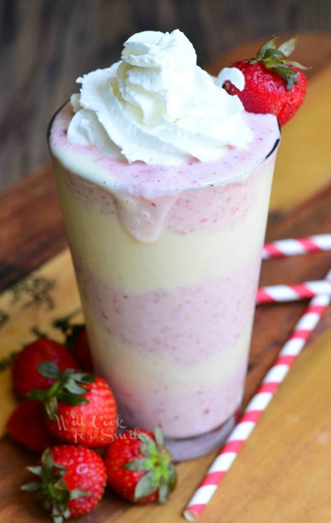 Strawberry Shortcake Milkshake layers of strawberry shake and cake in a tall glass cup with whipped cream and a strawberry as garnish