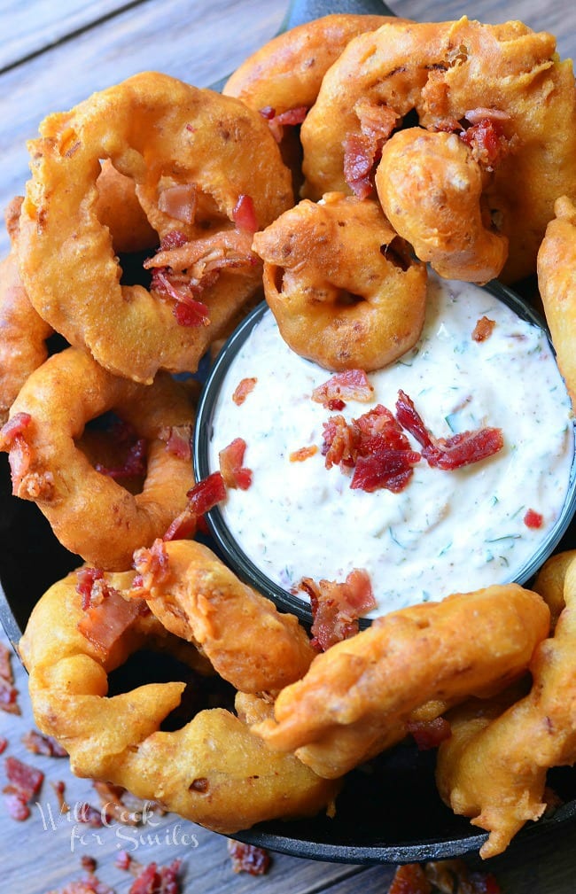 Top view of Bacon Onion Rings that are served in a black skillet around a dish of Bacon Ranch Dipping Sauce. Bacon is used to garnish the top of the dip.