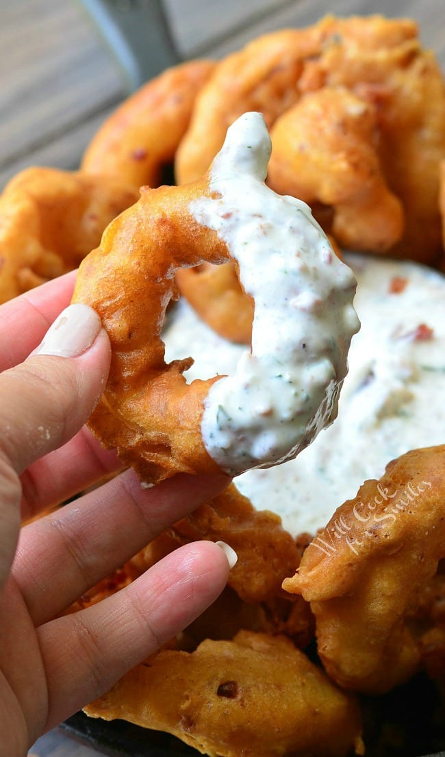 Bacon Onion Rings are served in a black skillet around a dish of Bacon Ranch Dipping Sauce, in the middle of the skillet. The left hand is holding an onion ring that has been dipped into the white sauce.