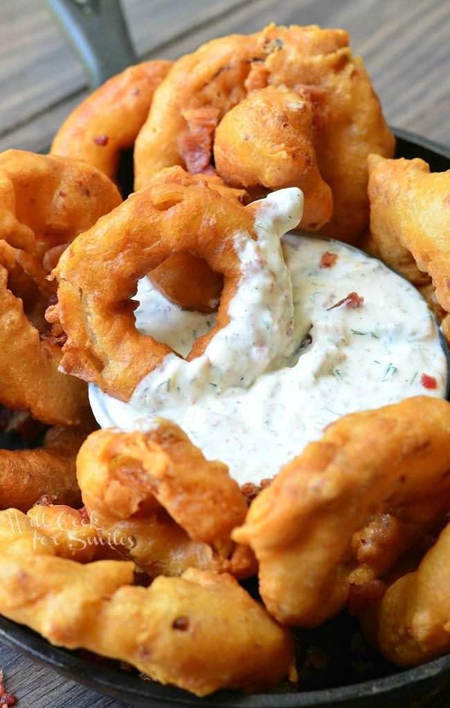 Bacon Onion Rings are served in a black skillet around a dish of Bacon Ranch Dipping Sauce, which is placed in the middle of the skillet. An onion ring that has been partially dipped into the sauce is laying on top of the dip.