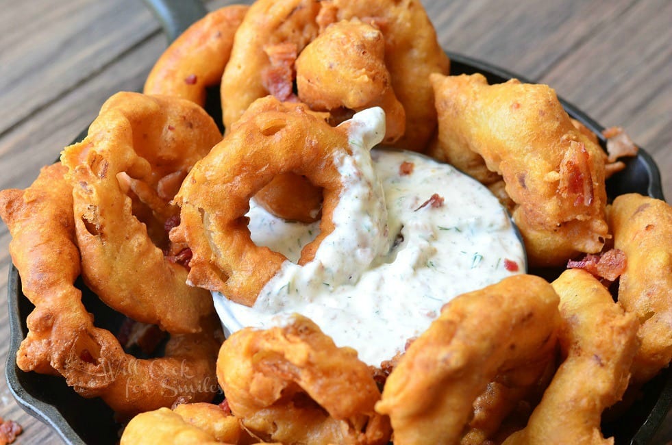 Bacon Onion Rings are served in a black skillet around a dish of Bacon Ranch Dipping Sauce, which is placed in the middle of the skillet. An onion ring that has been partially dipped into the sauce is laying on top of the dip.