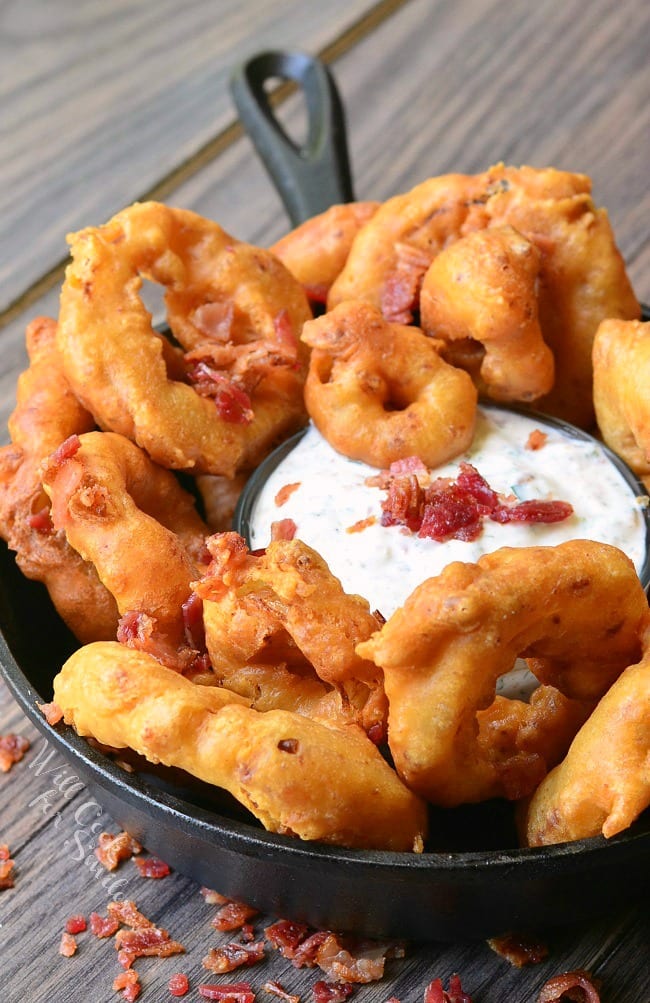 Bacon Onion Rings are served in a black skillet around a dish of Bacon Ranch Dipping Sauce. Bacon is used to garnish the top of the dip.