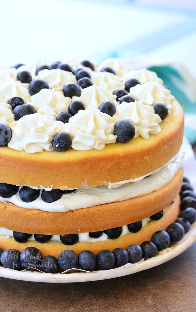 Blueberries and Cream Yellow Cake is a layered cake. There are three layers of yellow cake, white frosting, and blueberries. It's topped with more frosting and fresh blueberries. Around the border of the cake, on the plate, there are blueberries as well.