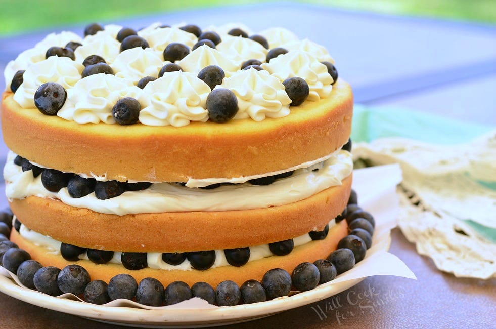 Side view of Blueberries and Cream Yellow Cake, which is a layered cake. There are three layers of yellow cake, white frosting, and blueberries. It's topped with more frosting and fresh blueberries. Around the border of the cake, on the plate, there are blueberries as well.