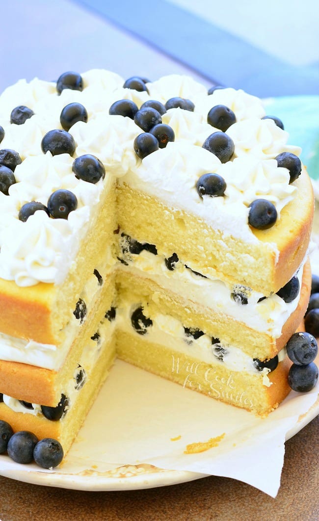 Blueberries and Cream Yellow Cake is a layered cake. There are three layers of yellow cake, white frosting, and blueberries. It's topped with more frosting and fresh blueberries. Around the border of the cake, on the plate, there are blueberries as well. One large piece of the cake has been cut and is missing from this view.