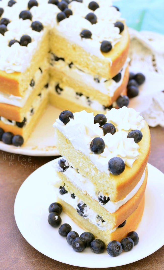 Blueberries and Cream Yellow Cake is a layered cake. There are three layers of yellow cake, white frosting, and blueberries. It's topped with more frosting and fresh blueberries. Around the border of the cake, on the plate, there are blueberries as well. One large piece of the cake has been cut and put on a separate plate. The rest of the cake is in the background.