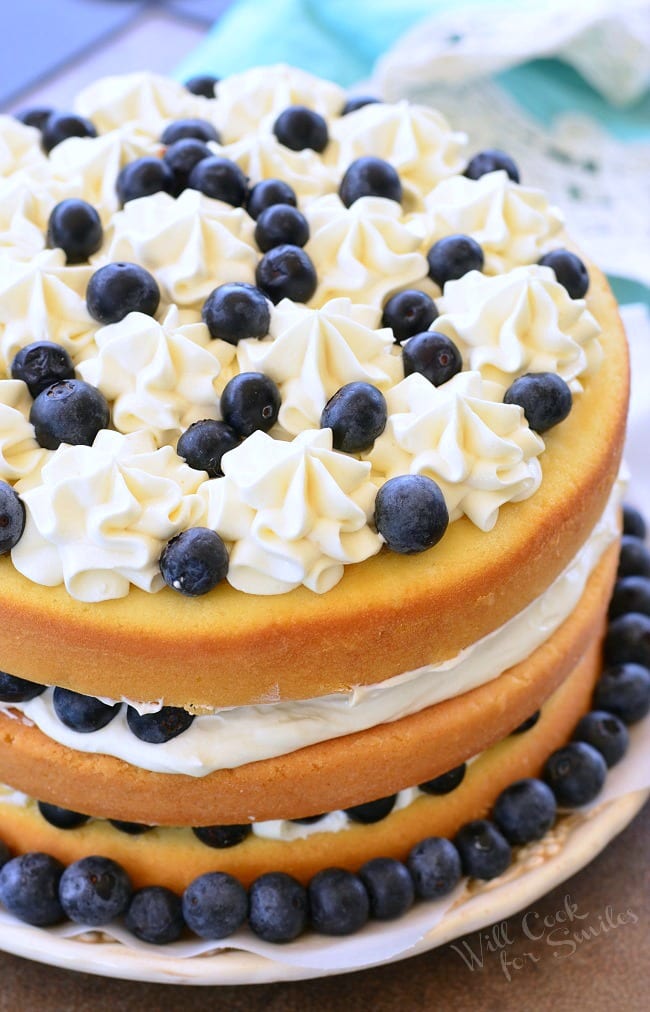 Blueberries and Cream Yellow Cake is a layered cake. There are three layers of yellow cake, white frosting, and blueberries. It's topped with more frosting and fresh blueberries. Around the border of the cake, on the plate, there are blueberries as well.