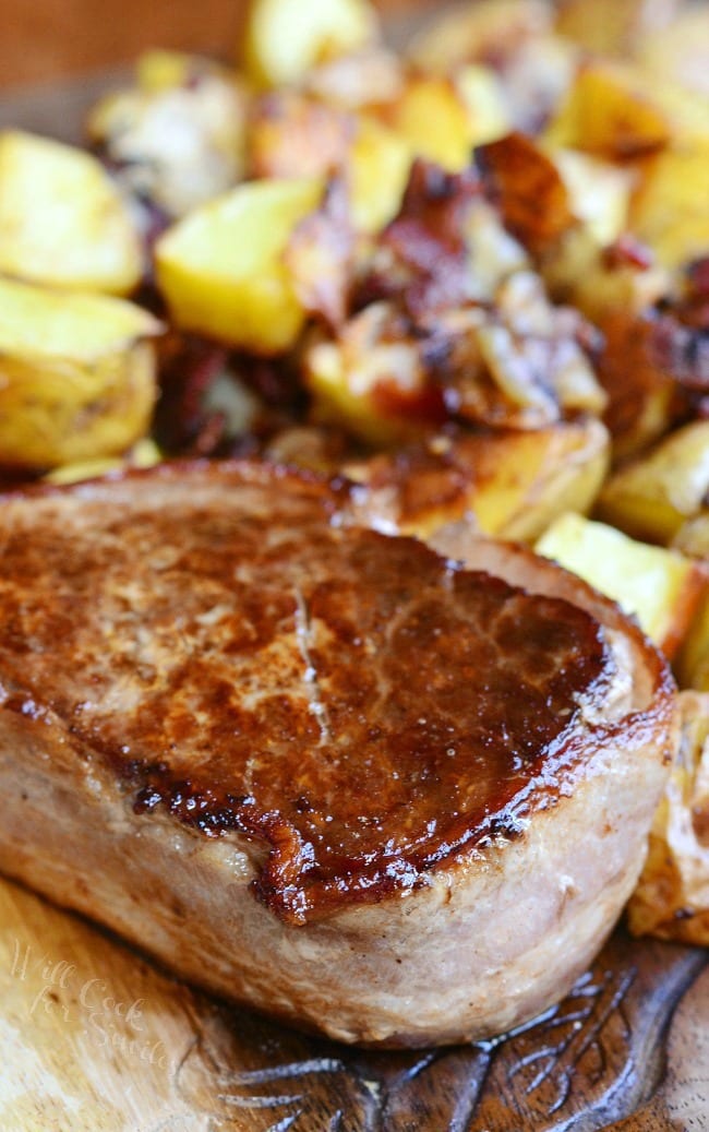 a cooked steak with a side of Roasted Potatoes with Bacon and Pearl Onions on a wood cutting board 