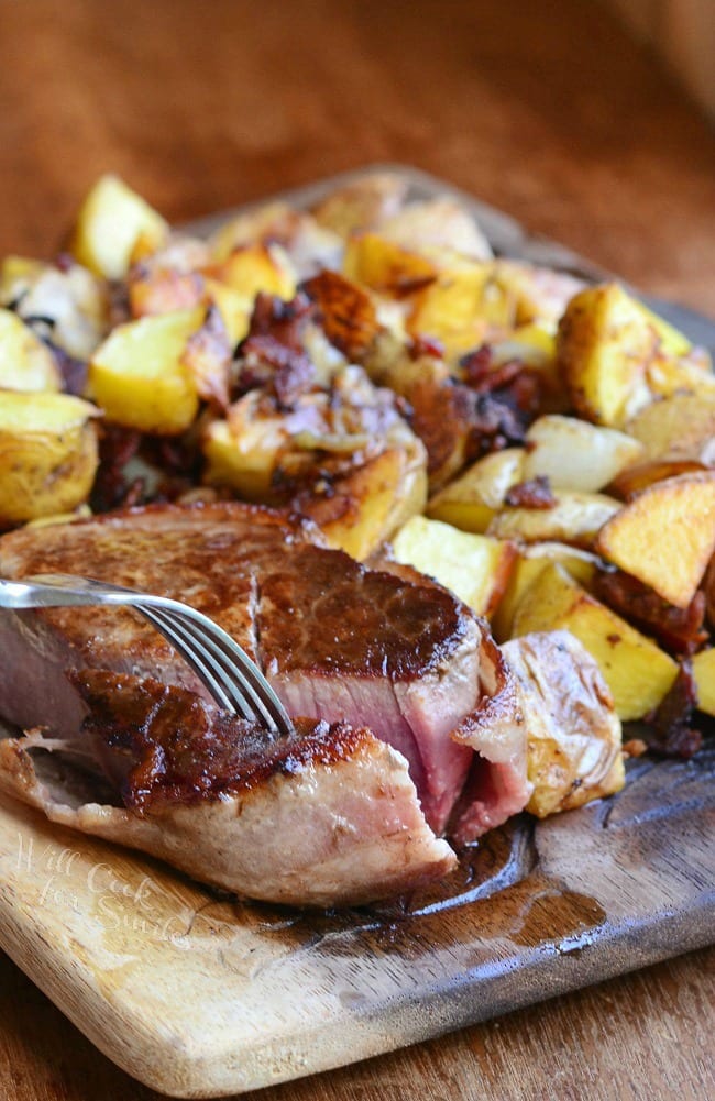 steak with a fork taking a piece and Roasted Potatoes with Bacon and Pearl Onions on a wood cutting board 