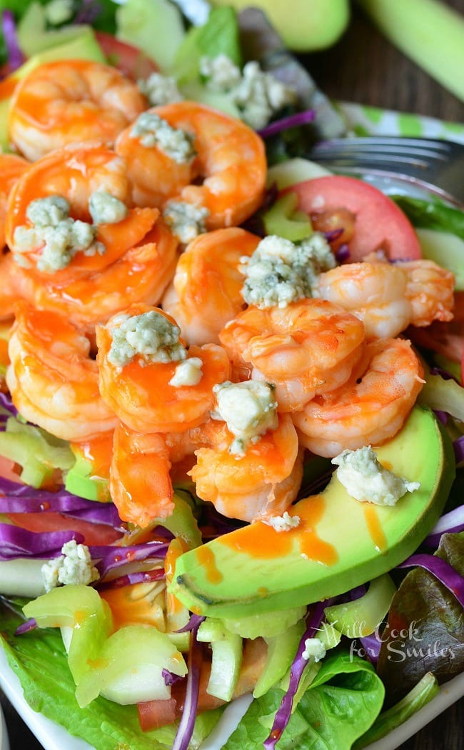 Close up of Buffalo Shrimp Salad with Homemade Blue Cheese Dressing that is presented on a white plate. On the bottom of the plate is greens. Purple cabbage and avocado slices are layered on top before the shrimp and blue cheese crumbles.
