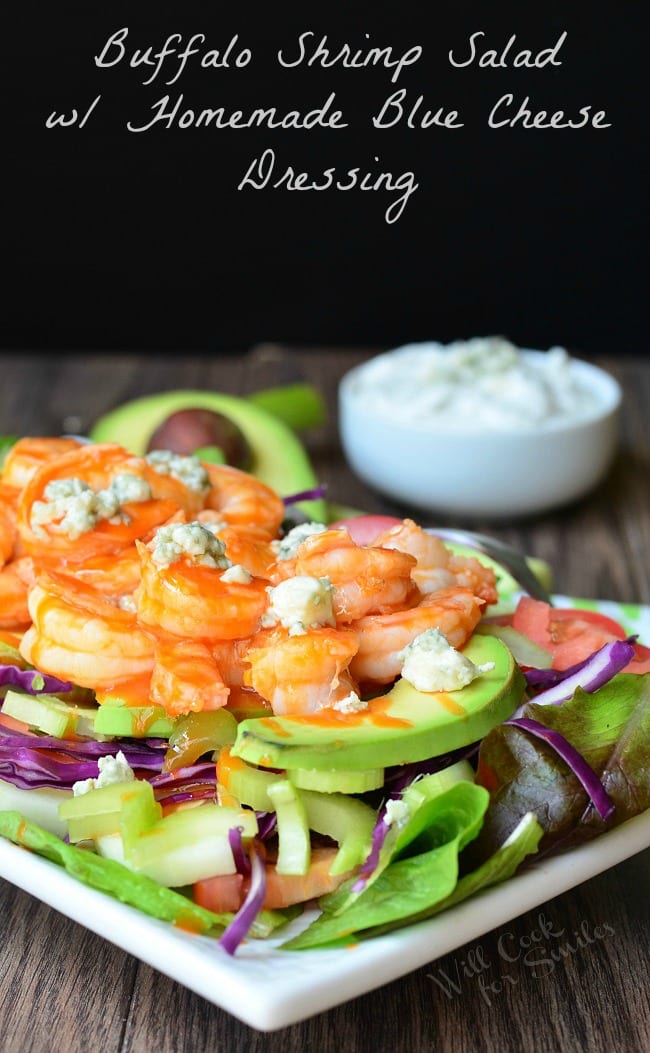 Buffalo Shrimp Salad with Homemade Blue Cheese Dressing is presented on a white plate. On the bottom of the plate is greens. Purple cabbage and avocado slices are layered on top before the shrimp and blue cheese crumbles. A half avocado and small bowl filled with dressing fade into the background.