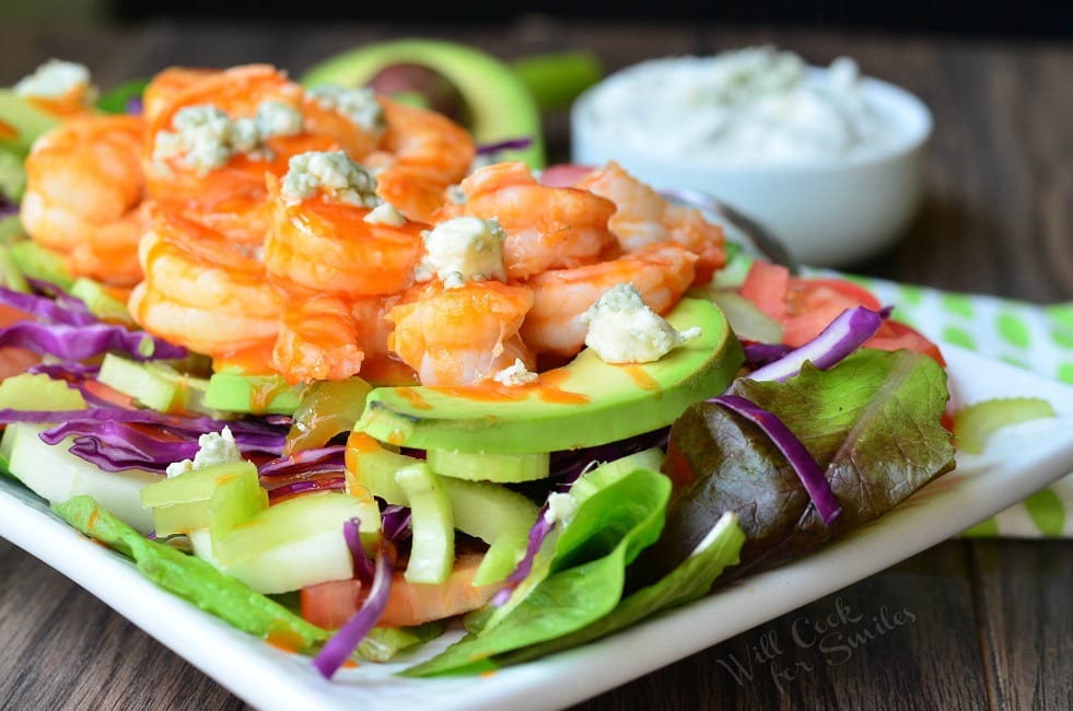 Buffalo Shrimp Salad with Homemade Blue Cheese Dressing is presented on a white plate. On the bottom of the plate is greens. Purple cabbage and avocado slices are layered on top before the shrimp and blue cheese crumbles. A half avocado and small bowl filled with dressing fade into the background.