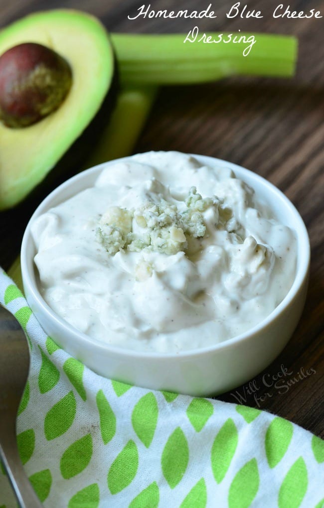 Homemade Blue Cheese Dressing is presented in a small, white bowl. It has blue cheese crumbles on top. Half an avocado is pictured in the background.