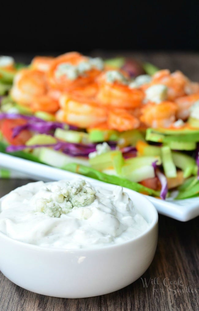 Homemade Blue Cheese Dressing is presented in a small, white bowl. It has blue cheese crumbles on top. In the background is the Buffalo Shrimp Salad.