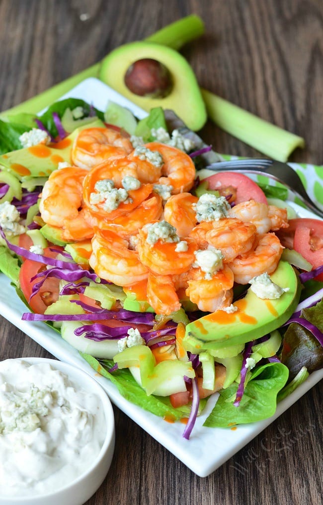 Top view of the Buffalo Shrimp Salad on a white plate. On the bottom of the plate is greens. Purple cabbage and avocado slices are layered on top before the shrimp and blue cheese crumbles. In the left front corner, the Homemade Blue Cheese Dressing is in a small, white bowl. It has blue cheese crumbles on top.  A half avocado and celery sticks are in the background.