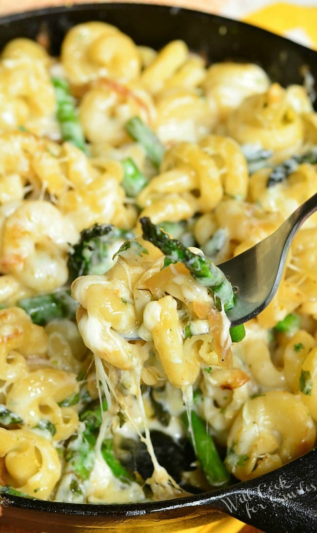 Extra Cheesy Lemon Asparagus Pasta Skillet is presented here in a black skillet. In the skillet, there are many noodles and asparagus covered in a creamy sauce. A fork is above the skillet holding some of the pasta up.