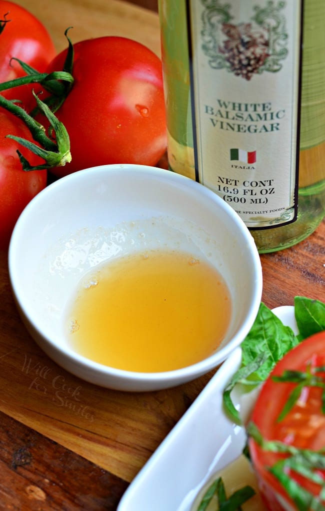 There is a white bowl with white balsamic reduction in it. It's yellow in color. There is a bottle of white balsamic vinegar and whole tomatoes in the background.