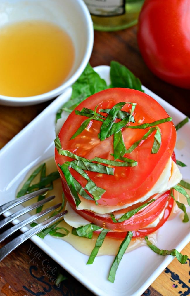 Top view of Fresh Caprese Salad with White Balsamic Reduction is in a small, white serving dish with a fork. There is stacked tomatoes, mozzarella cheese, and chopped basil leaves. There is a white bowl with extra white balsamic reduction in it and whole tomatoes in the background.