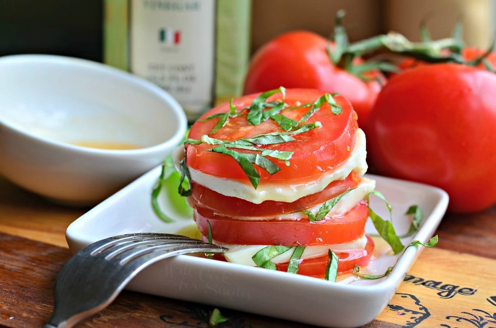 Side view of Fresh Caprese Salad with White Balsamic Reduction is in a small, white serving dish with a fork. There is stacked tomatoes, mozzarella cheese, and chopped basil leaves. There is a white bowl with extra white balsamic reduction in it and whole tomatoes in the background.