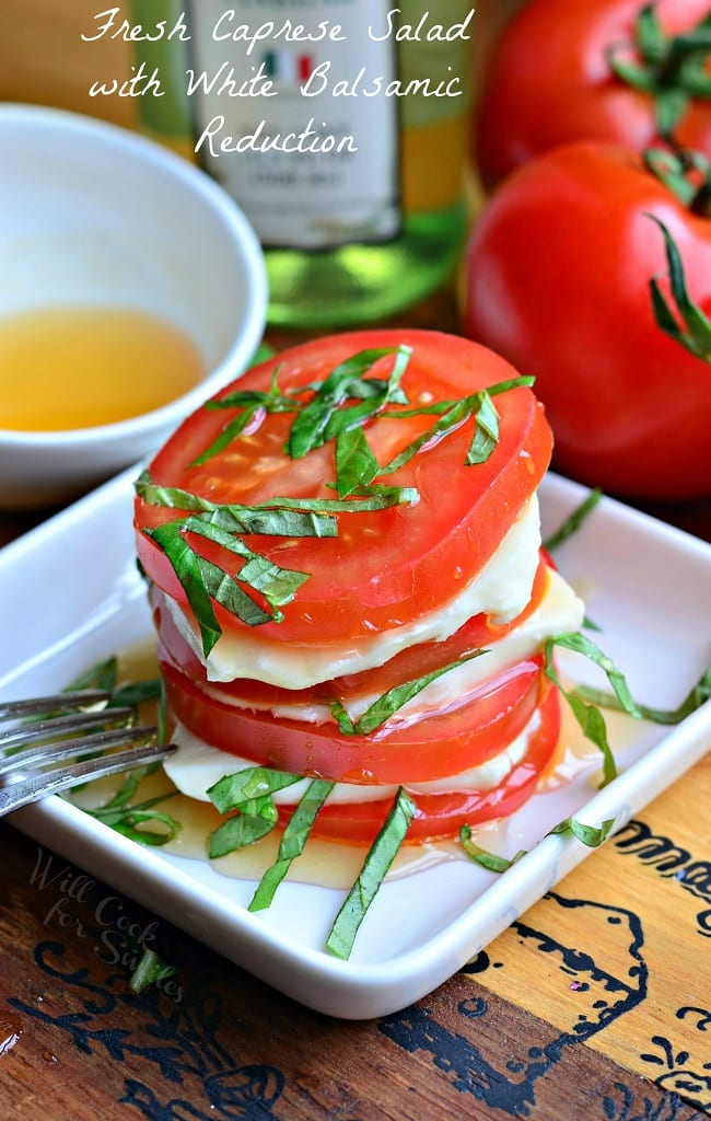 Fresh Caprese Salad with White Balsamic Reduction is in a small, white serving dish with a fork. There is stacked tomatoes, mozzarella cheese, and chopped basil leaves. There is a white bowl with extra white balsamic reduction in it and whole tomatoes in the background.