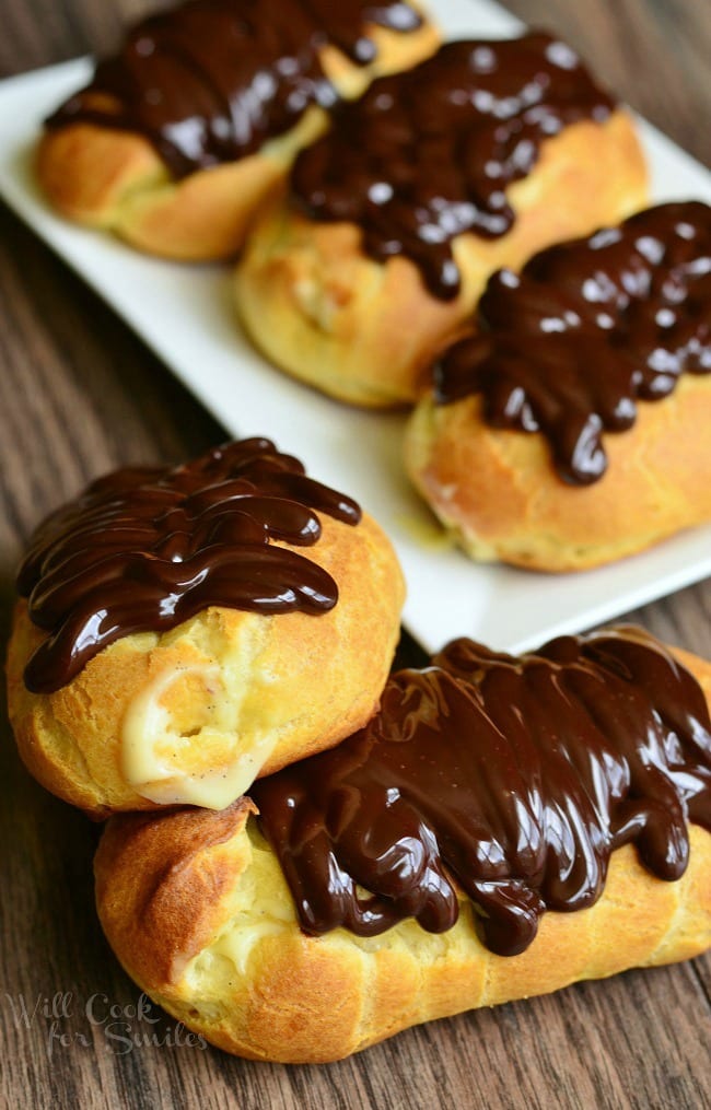 Homemade Boston Cream Eclairs are a golden, brown color. Two sit in front on a wooden surface. They are filled with homemade vanilla bean custard and topped with rich chocolate ganache. More Eclairs lay on a white plate in the background.