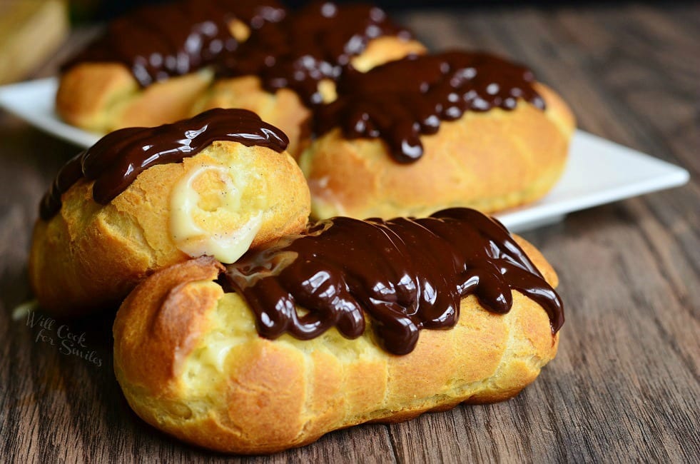 Homemade Boston Cream Eclairs are a golden, brown color. Two sit in front on a wooden surface. They are filled with homemade vanilla bean custard and topped with rich chocolate ganache. More Eclairs lay on a white plate in the background.