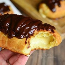 hand holding up one eclair with a bite in it above wooden table and more eclairs on a wooden cutting board