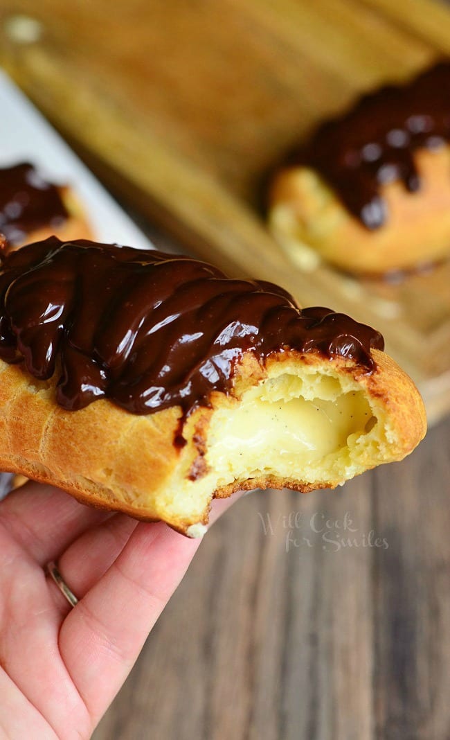 A hand holds a Homemade Boston Cream Eclair. A bite has been taken out of the Eclair. The pastry is a golden, brown color. They are filled with homemade vanilla bean custard and topped with rich chocolate ganache.