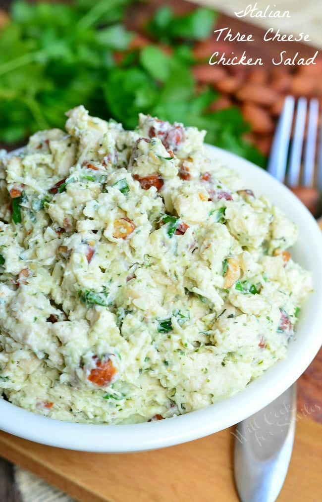 white bowl filled with italian three cheese chicken salad on a wooden cutting board with a fork below the rim of the bowl to the right
