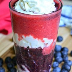 Layered berry daiquiri red white and blue smoothie on a wooden cutting board with blue berries scattered around base of glass