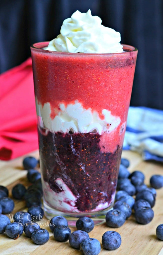 Layered Berry Daiquiri Red White and Blue Daiquiri is in a tall glass. There are layers of whipped cream and berries up to the rim. The top has whipped cream as well. Blueberries are scattered around the glass on a wooden board.